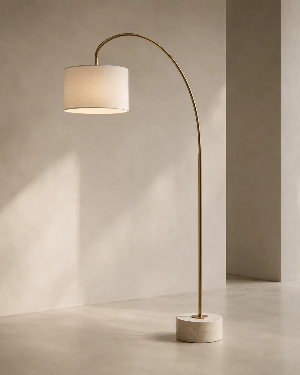 Brushed brass arc floor lamp with a linen drum shade over a stone-effect base in a sunlit studio setting